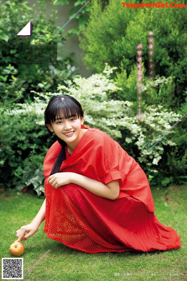 A woman in a red dress sitting on the grass with an apple.